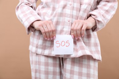 Photo of Woman holding sticky note with word SOS and suffering from cystitis symptoms on dark beige background, closeup