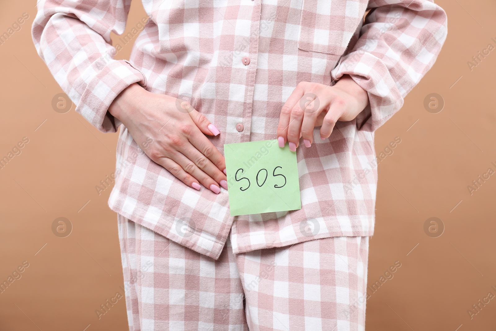 Photo of Woman holding sticky note with word SOS and suffering from cystitis symptoms on dark beige background, closeup