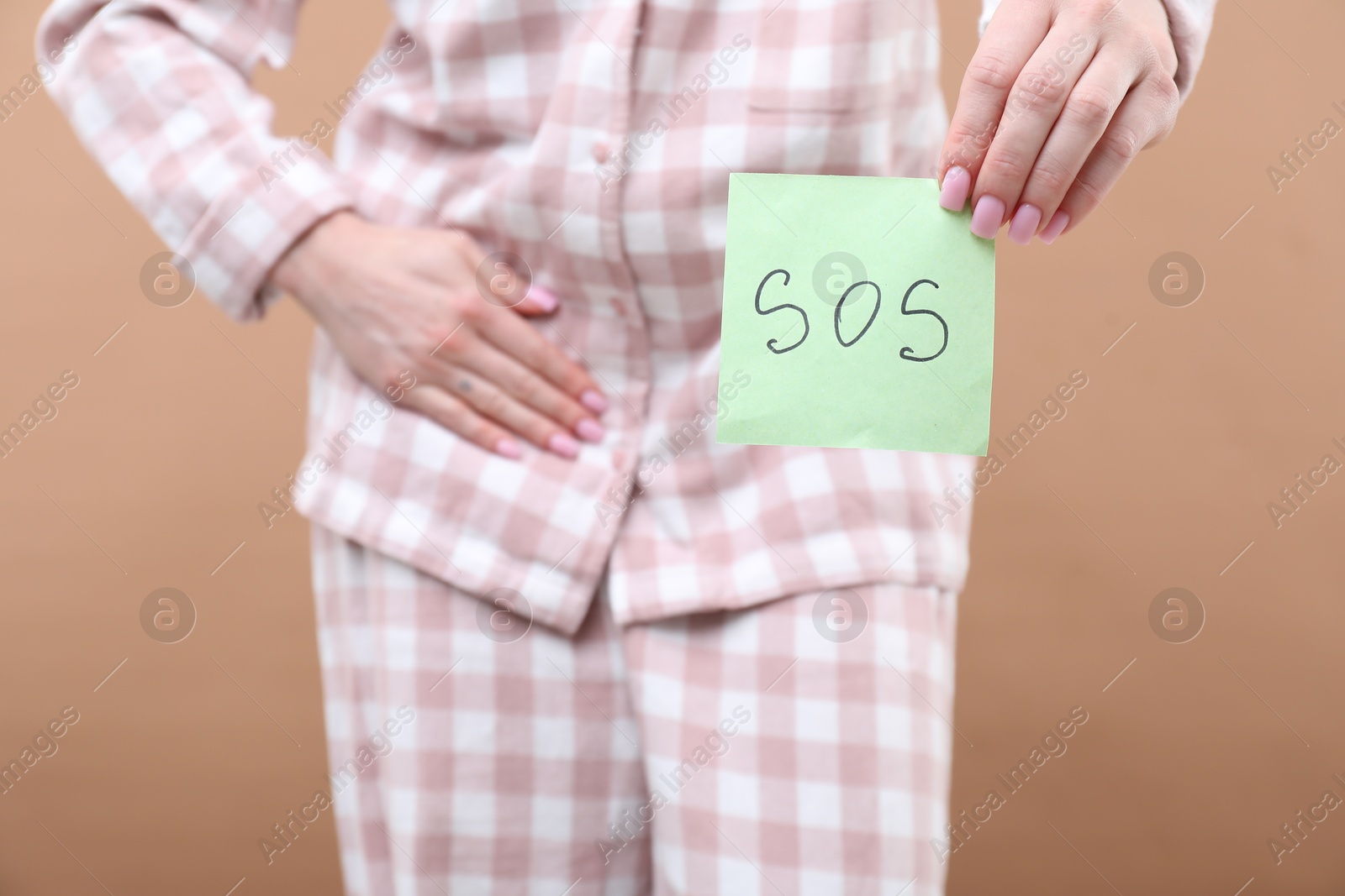 Photo of Woman holding sticky note with word SOS and suffering from cystitis symptoms on dark beige background, closeup