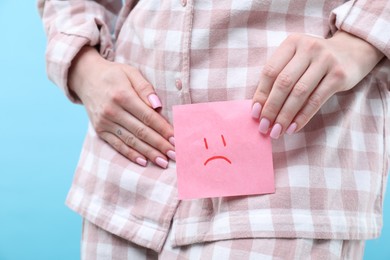 Photo of Woman holding sticky note with drawn sad face and suffering from cystitis symptoms on light blue background, closeup