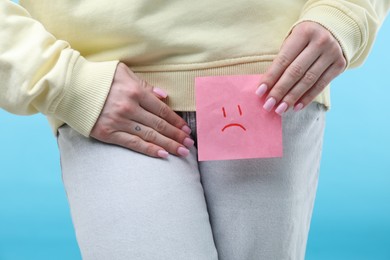 Photo of Woman holding sticky note with drawn sad face and suffering from cystitis symptoms on light blue background, closeup