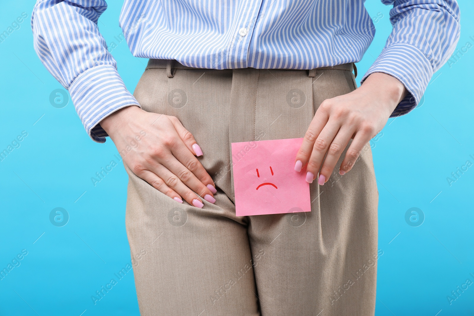 Photo of Woman holding sticky note with drawn sad face and suffering from cystitis symptoms on light blue background, closeup