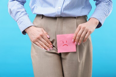 Photo of Woman holding sticky note with drawn sad face and suffering from cystitis symptoms on light blue background, closeup