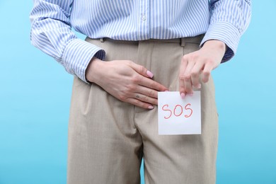 Photo of Woman holding sticky note with word SOS and suffering from cystitis symptoms on light blue background, closeup