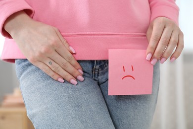 Photo of Woman holding sticky note with drawn sad face and suffering from cystitis symptoms at home, closeup