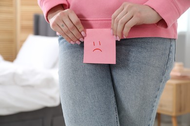 Photo of Woman holding sticky note with drawn sad face and suffering from cystitis symptoms at home, closeup
