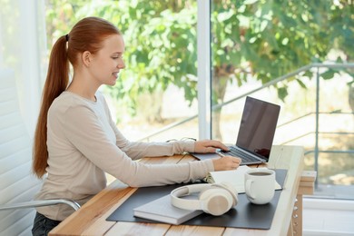 Photo of Teenage girl using laptop at wooden desk indoors. Home workplace