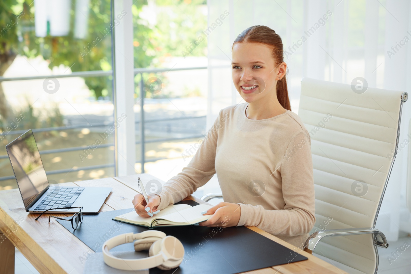 Photo of Teenage girl writing in notebook at wooden desk indoors. Home workplace