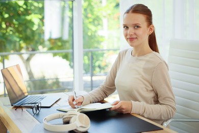 Photo of Teenage girl writing in notebook at wooden desk indoors. Home workplace