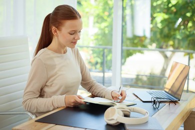Photo of Teenage girl writing in notebook at wooden desk indoors. Home workplace