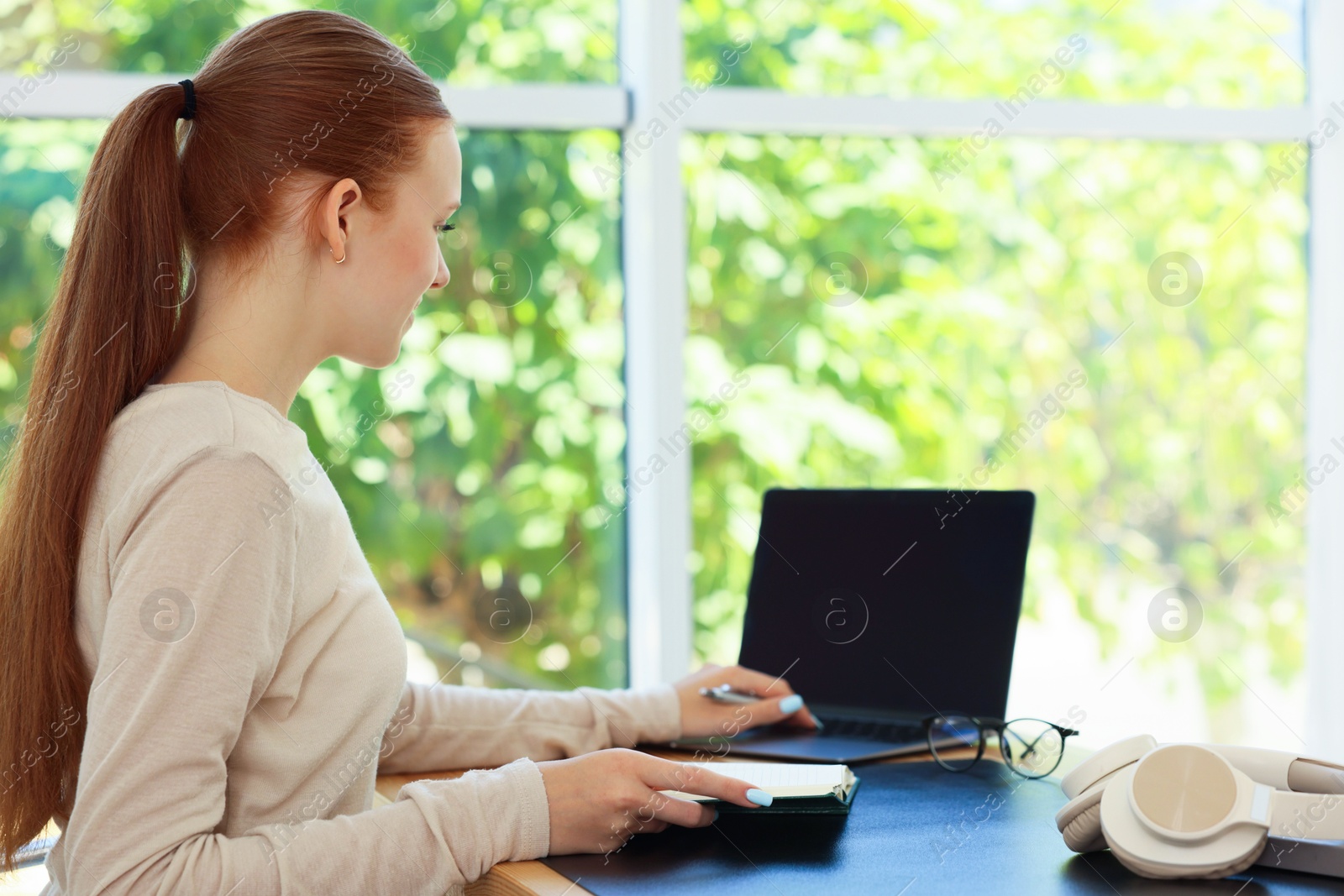 Photo of Teenage girl with book using laptop at wooden desk indoors. Home workplace