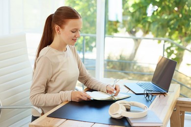 Photo of Teenage girl writing in notebook and using laptop at wooden desk indoors. Home workplace
