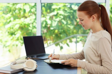 Photo of Teenage girl writing in notebook and using laptop at wooden desk indoors. Home workplace