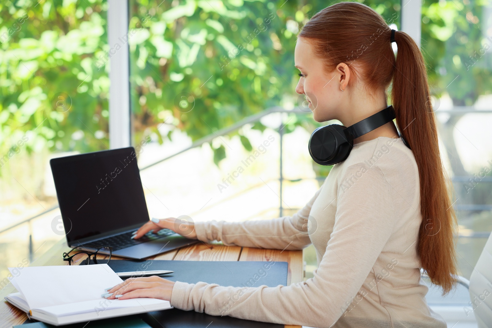 Photo of Teenage girl with book using laptop at wooden desk indoors. Home workplace