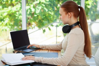 Photo of Teenage girl with book using laptop at wooden desk indoors. Home workplace