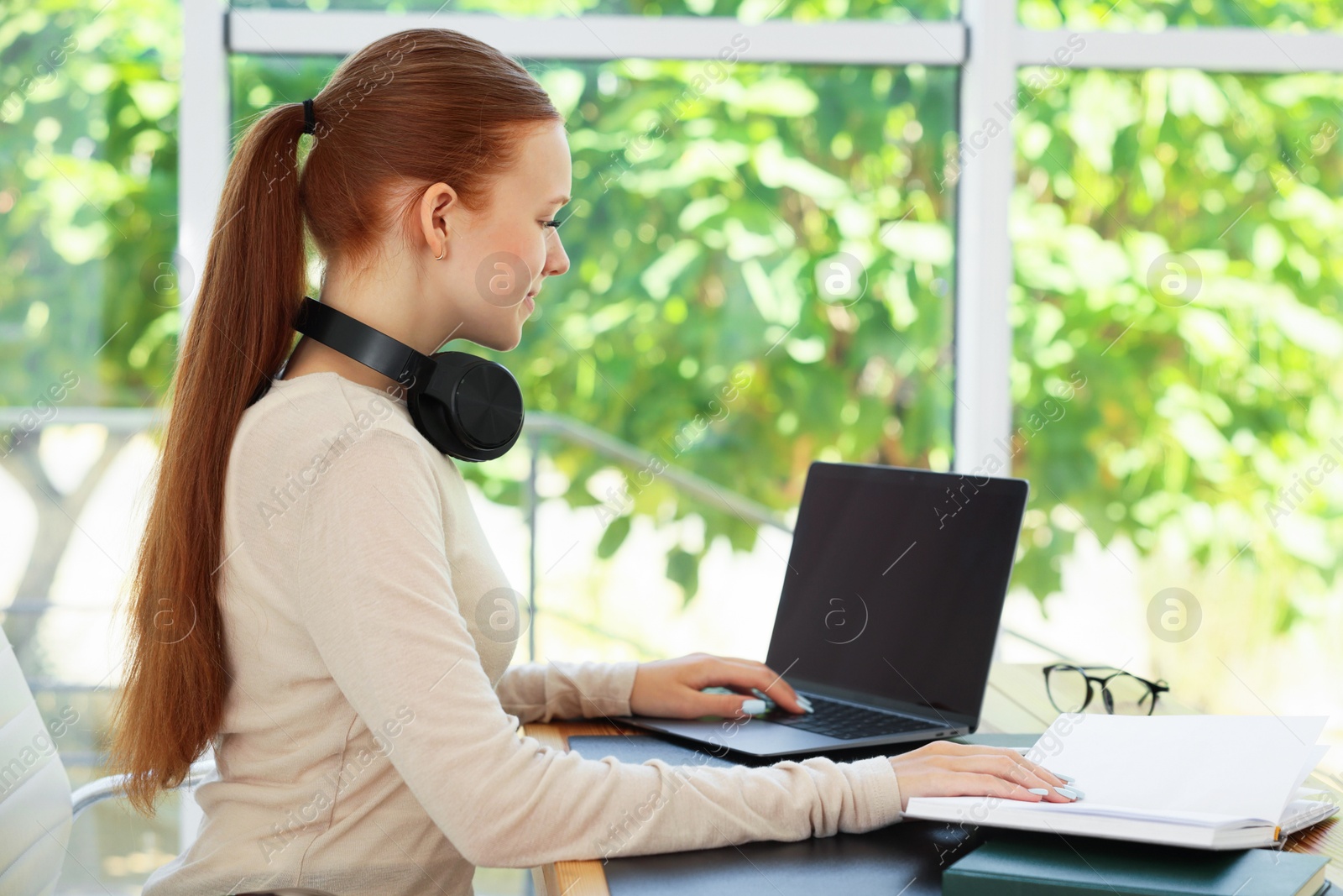 Photo of Teenage girl with book using laptop at wooden desk indoors. Home workplace