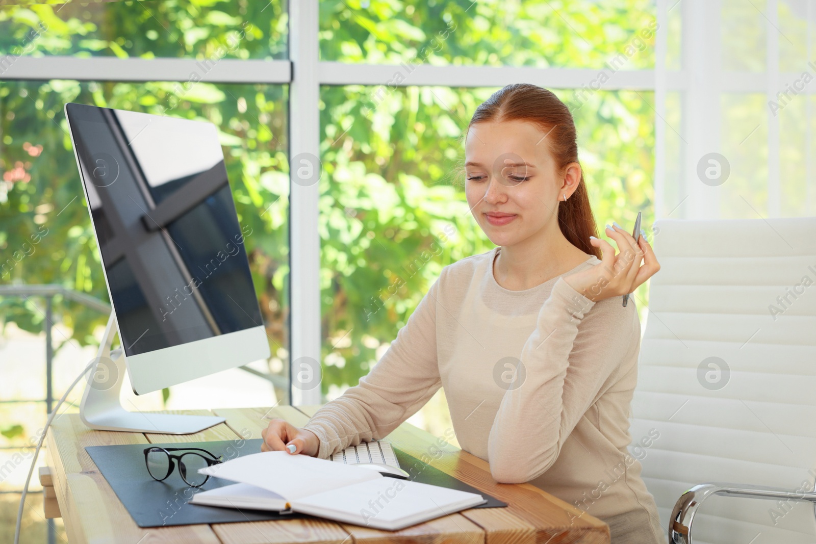 Photo of Teenage girl with book using computer at wooden desk indoors. Home workplace