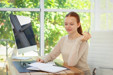 Photo of Teenage girl with book using computer at wooden desk indoors. Home workplace