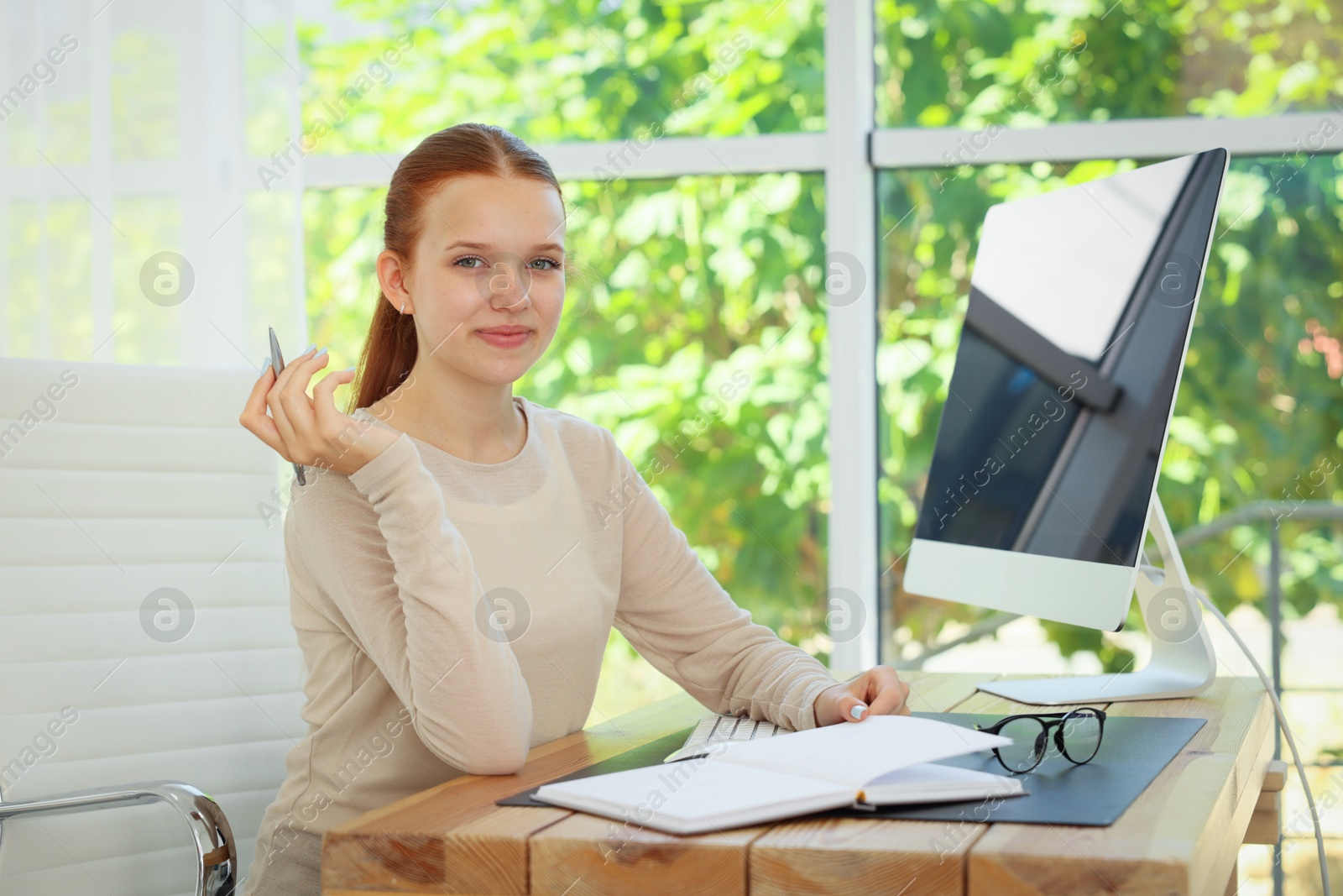 Photo of Teenage girl with book using computer at wooden desk indoors. Home workplace