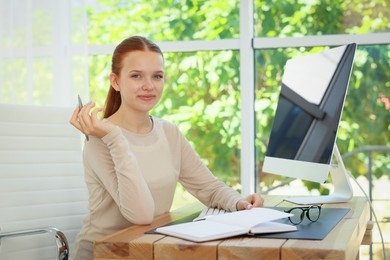 Photo of Teenage girl with book using computer at wooden desk indoors. Home workplace