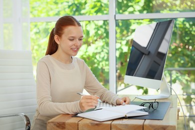Photo of Teenage girl using computer and writing in notebook at wooden desk indoors. Home workplace