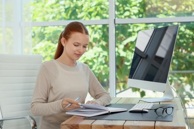 Photo of Teenage girl with book using computer at wooden desk indoors. Home workplace