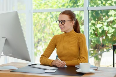 Photo of Teenage girl using computer and writing in notebook at wooden desk indoors. Home workplace