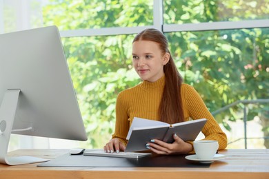 Photo of Teenage girl with book using computer at wooden desk indoors. Home workplace