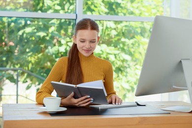 Photo of Teenage girl with book using computer at wooden desk indoors. Home workplace