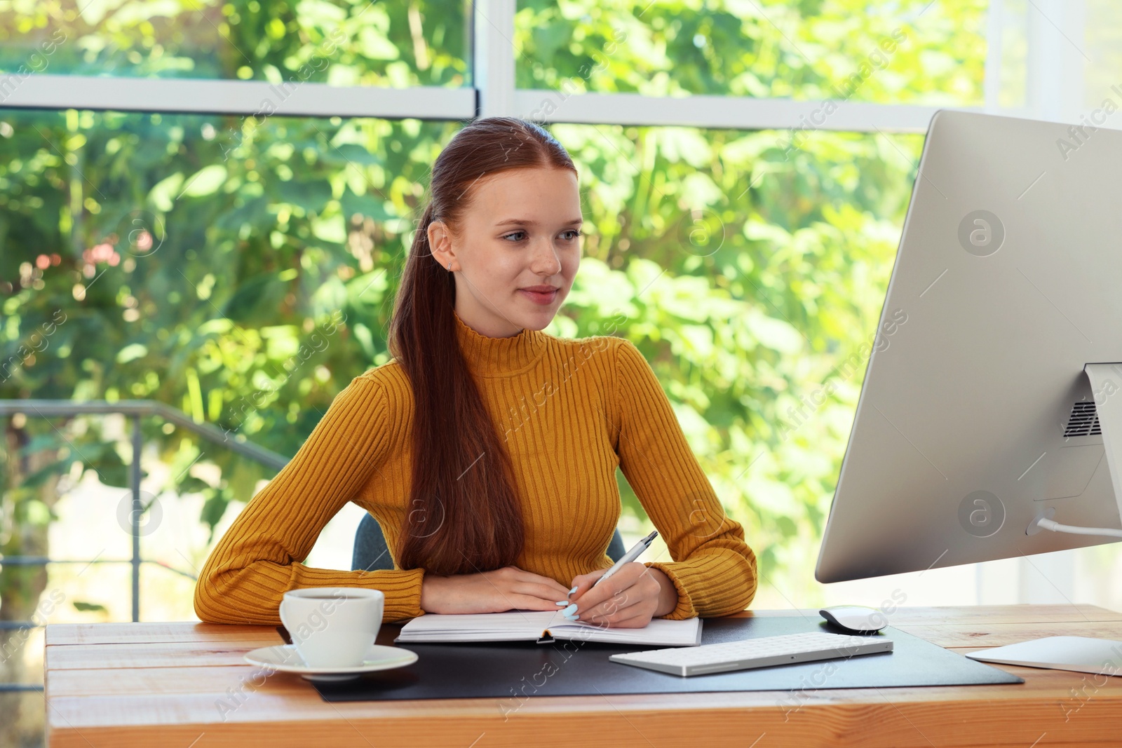 Photo of Teenage girl using computer and writing in notebook at wooden desk indoors. Home workplace