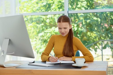 Photo of Teenage girl using computer and writing in notebook at wooden desk indoors. Home workplace
