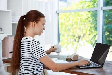 Photo of Teenage girl with cup of drink using laptop at wooden desk indoors. Home workplace