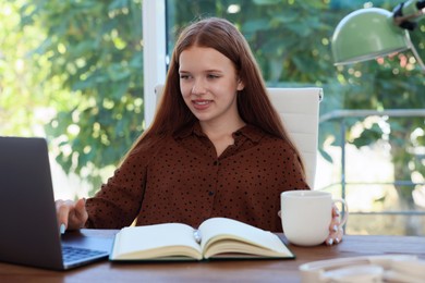 Photo of Teenage girl with cup of drink using laptop at wooden desk indoors. Home workplace