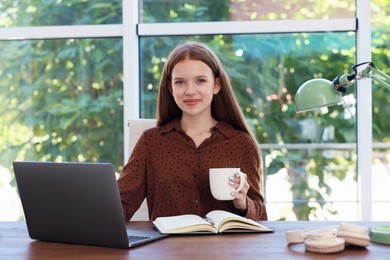 Photo of Teenage girl with cup of drink using laptop at wooden desk indoors. Home workplace