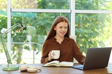 Photo of Teenage girl with cup of drink using laptop at wooden desk indoors. Home workplace
