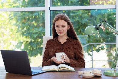 Photo of Teenage girl with cup of drink using laptop at wooden desk indoors. Home workplace