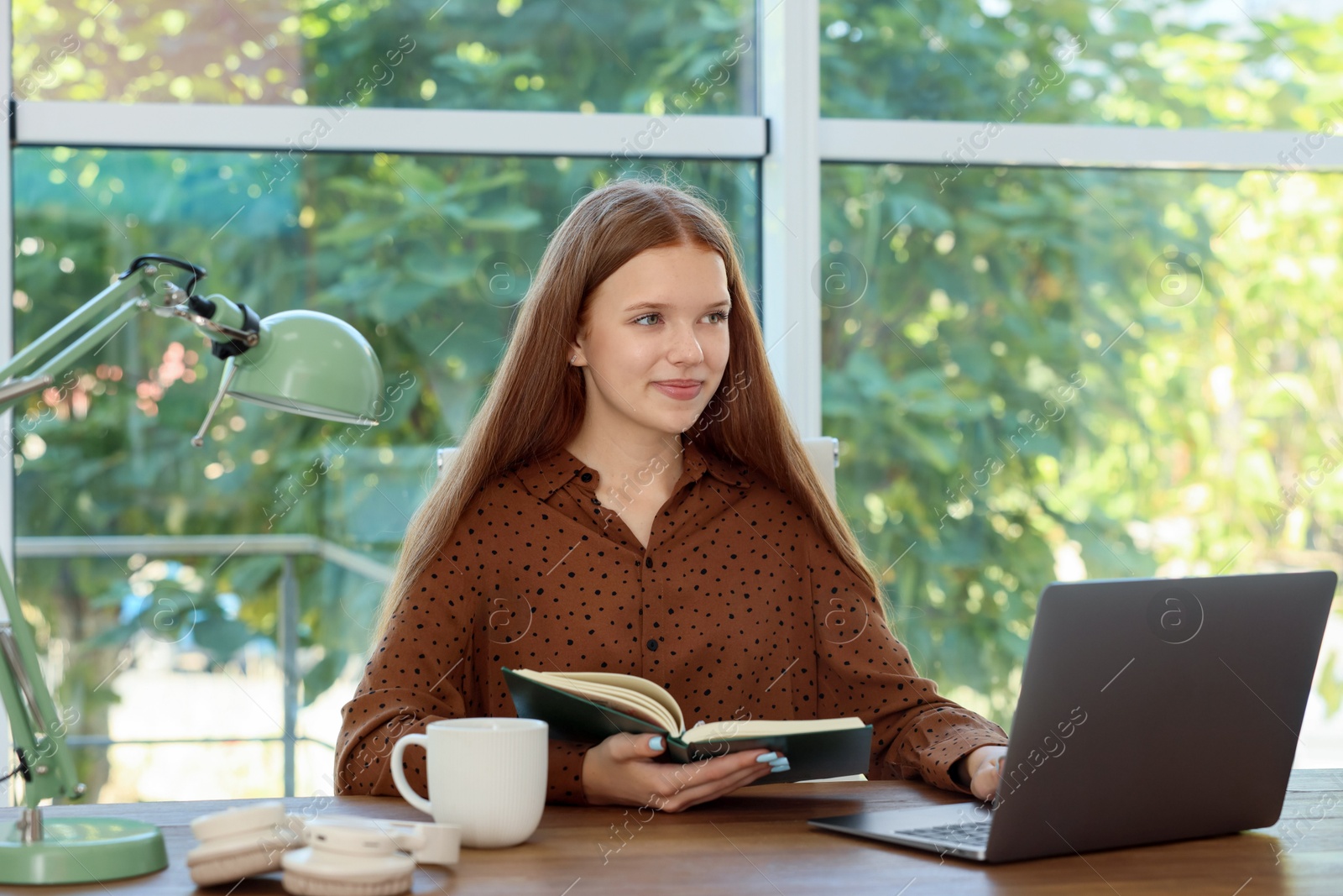 Photo of Teenage girl with book using laptop at wooden desk indoors. Home workplace