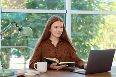 Photo of Teenage girl with book using laptop at wooden desk indoors. Home workplace