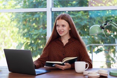 Photo of Teenage girl with book using laptop at wooden desk indoors. Home workplace