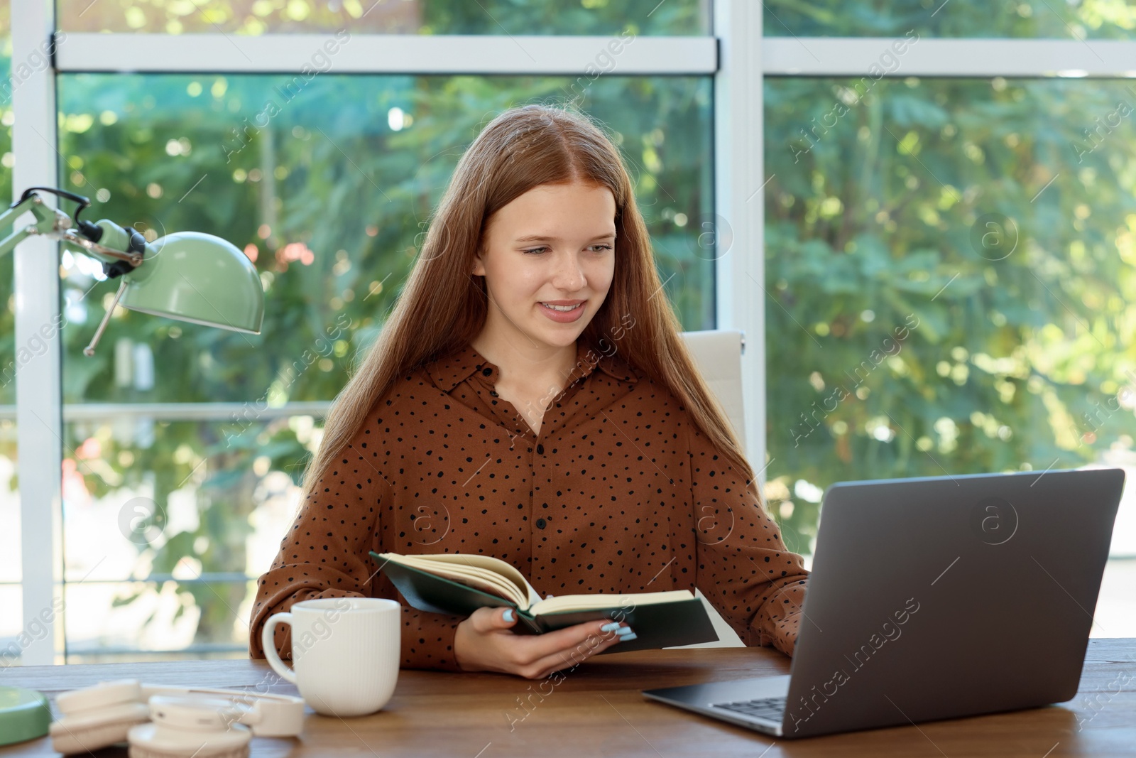 Photo of Teenage girl with book using laptop at wooden desk indoors. Home workplace