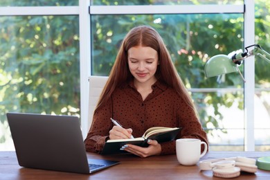 Photo of Teenage girl using laptop and writing in notebook at wooden desk indoors. Home workplace