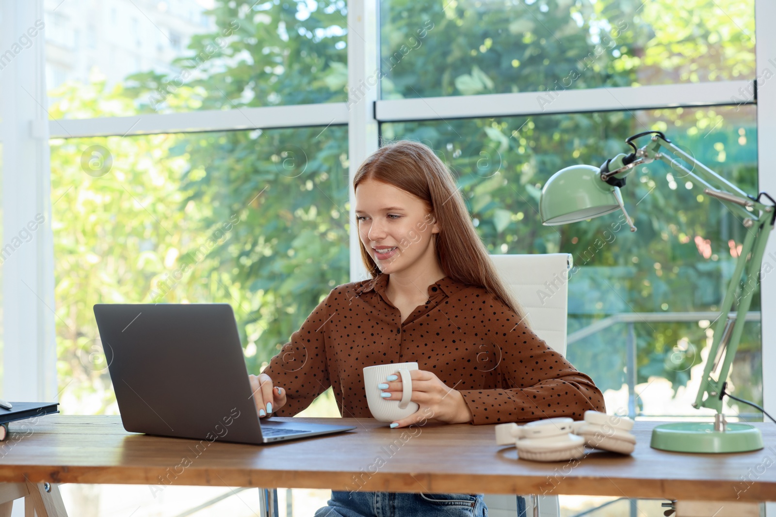 Photo of Teenage girl with cup of drink using laptop at wooden desk indoors. Home workplace