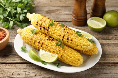 Photo of Tasty grilled corncobs with parsley and lime slices on wooden table, closeup