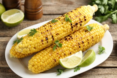 Photo of Tasty grilled corncobs with parsley and lime slices on wooden table, closeup