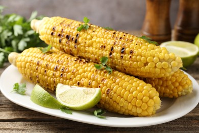 Photo of Tasty grilled corncobs with parsley and lime slices on wooden table, closeup