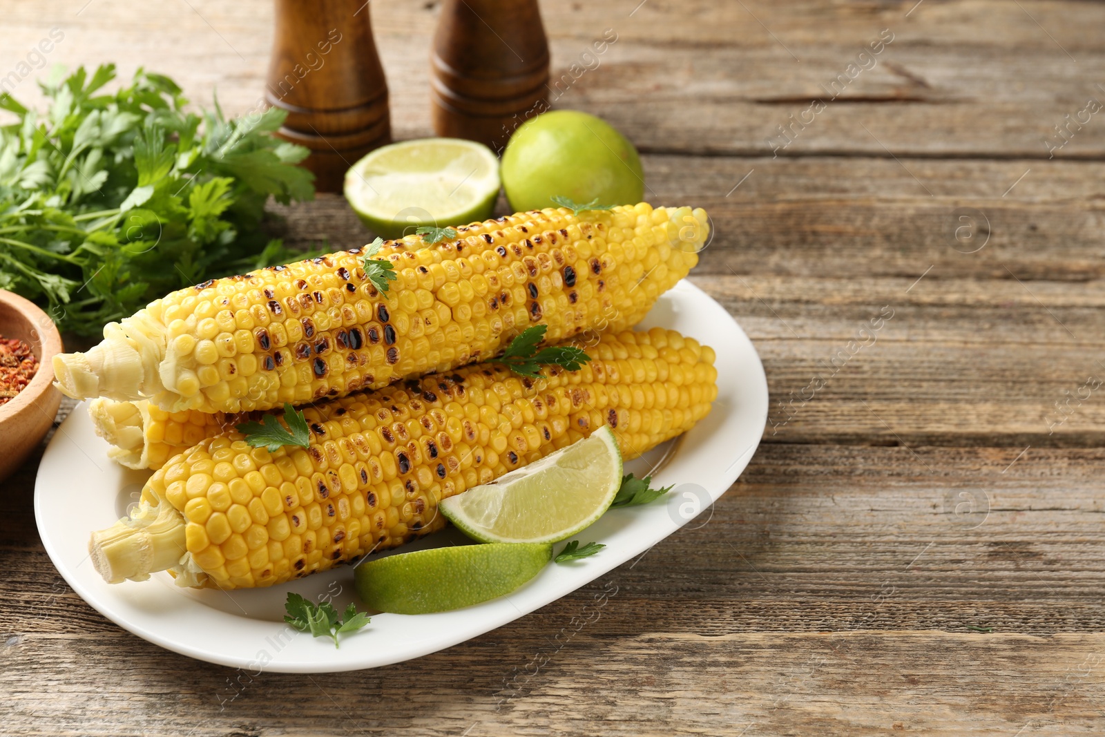 Photo of Tasty grilled corncobs with parsley and lime slices on wooden table. Space for text