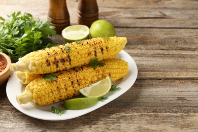 Photo of Tasty grilled corncobs with parsley and lime slices on wooden table. Space for text