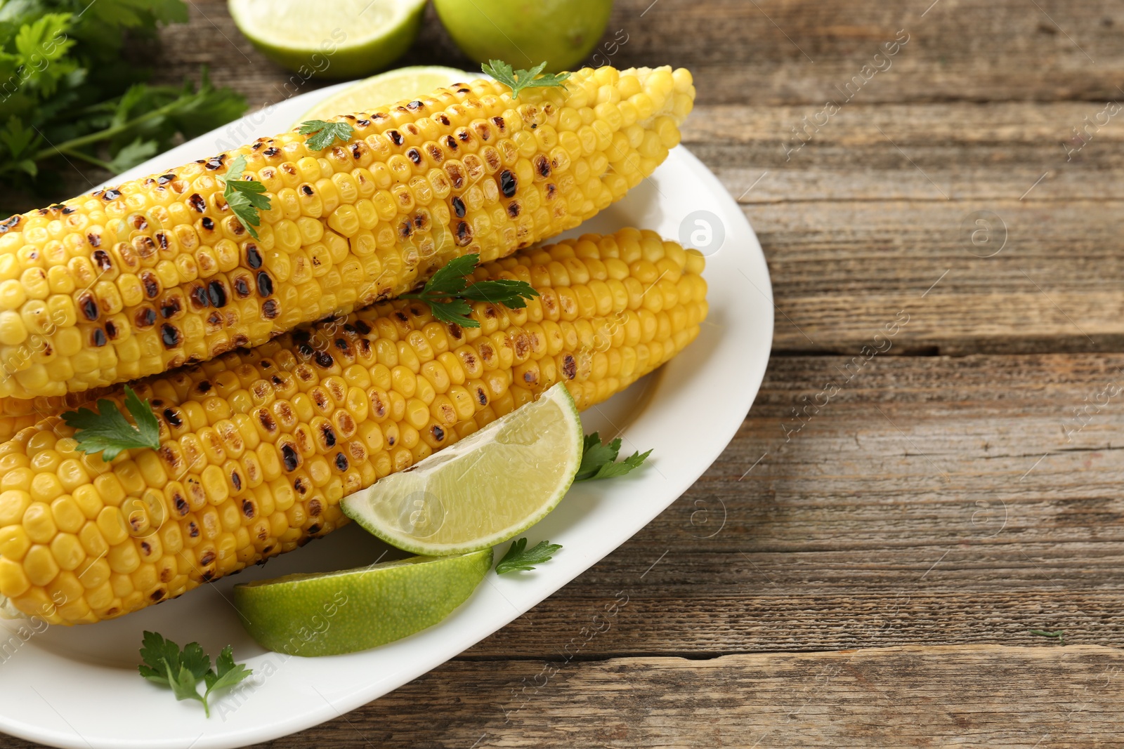 Photo of Tasty grilled corncobs with parsley and lime slices on wooden table, closeup. Space for text