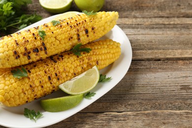 Photo of Tasty grilled corncobs with parsley and lime slices on wooden table, closeup. Space for text