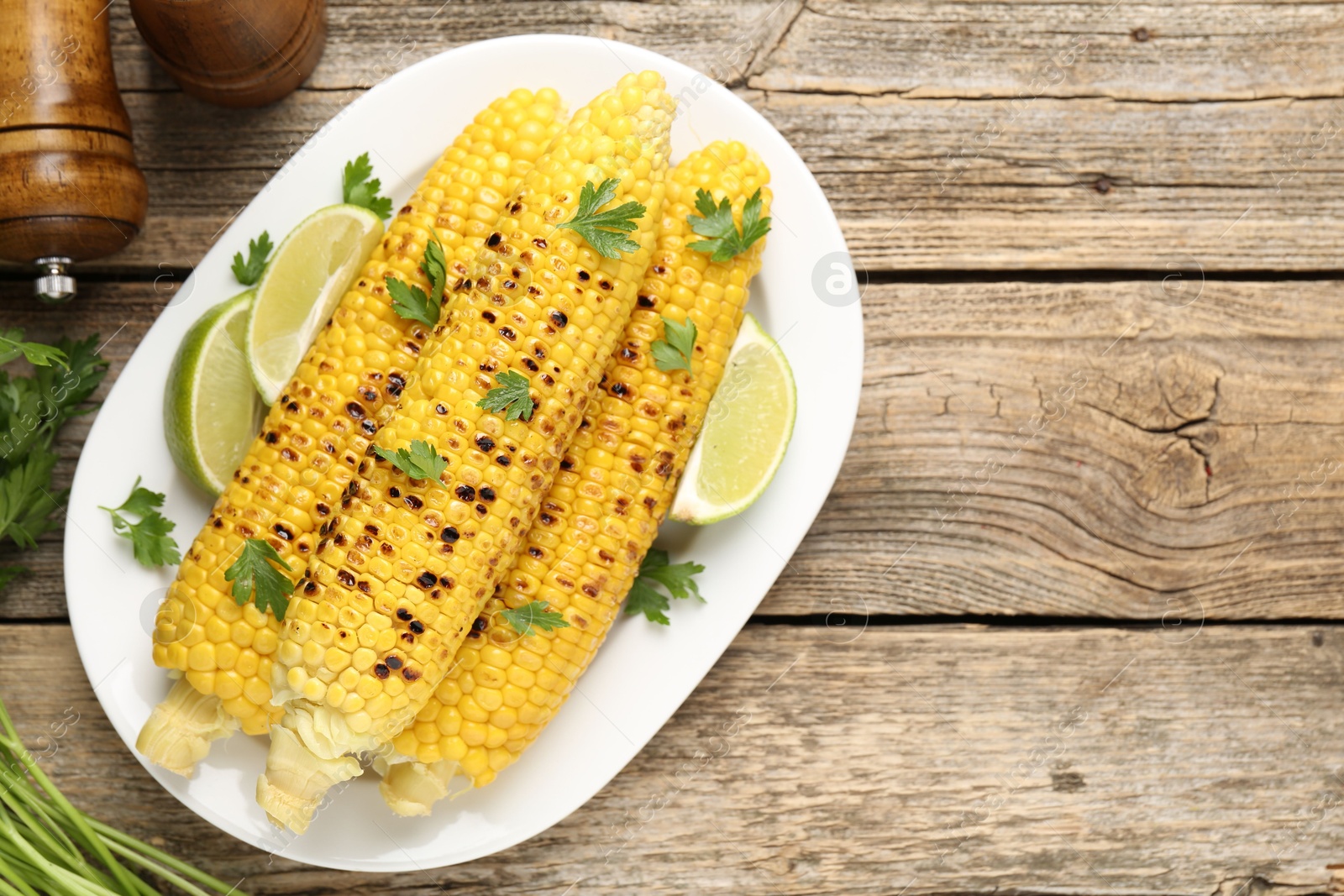 Photo of Tasty grilled corncobs with parsley and lime slices on wooden table, flat lay. Space for text